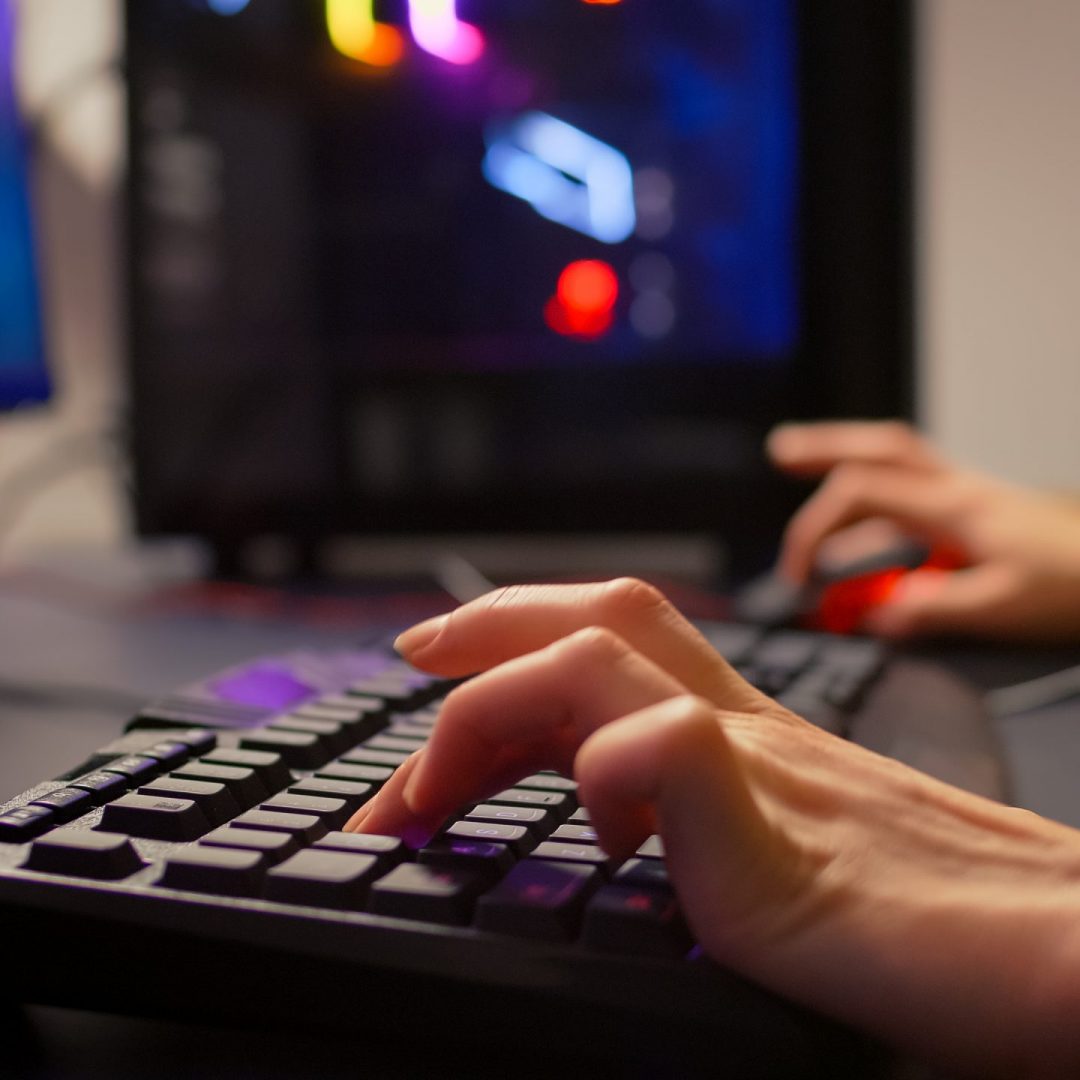 Close-up of gamer using RGB keyboard and mouse for online competition. Professional gamer playing space shooter video games in gamming room with neon light